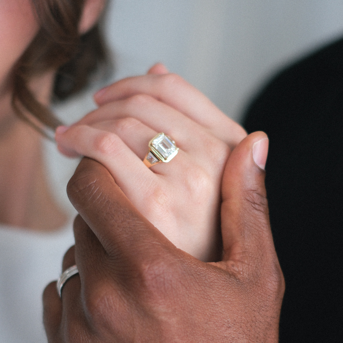 Close-up of two hands holding each other, one with a gold ring featuring an emerald-cut gemstone.
